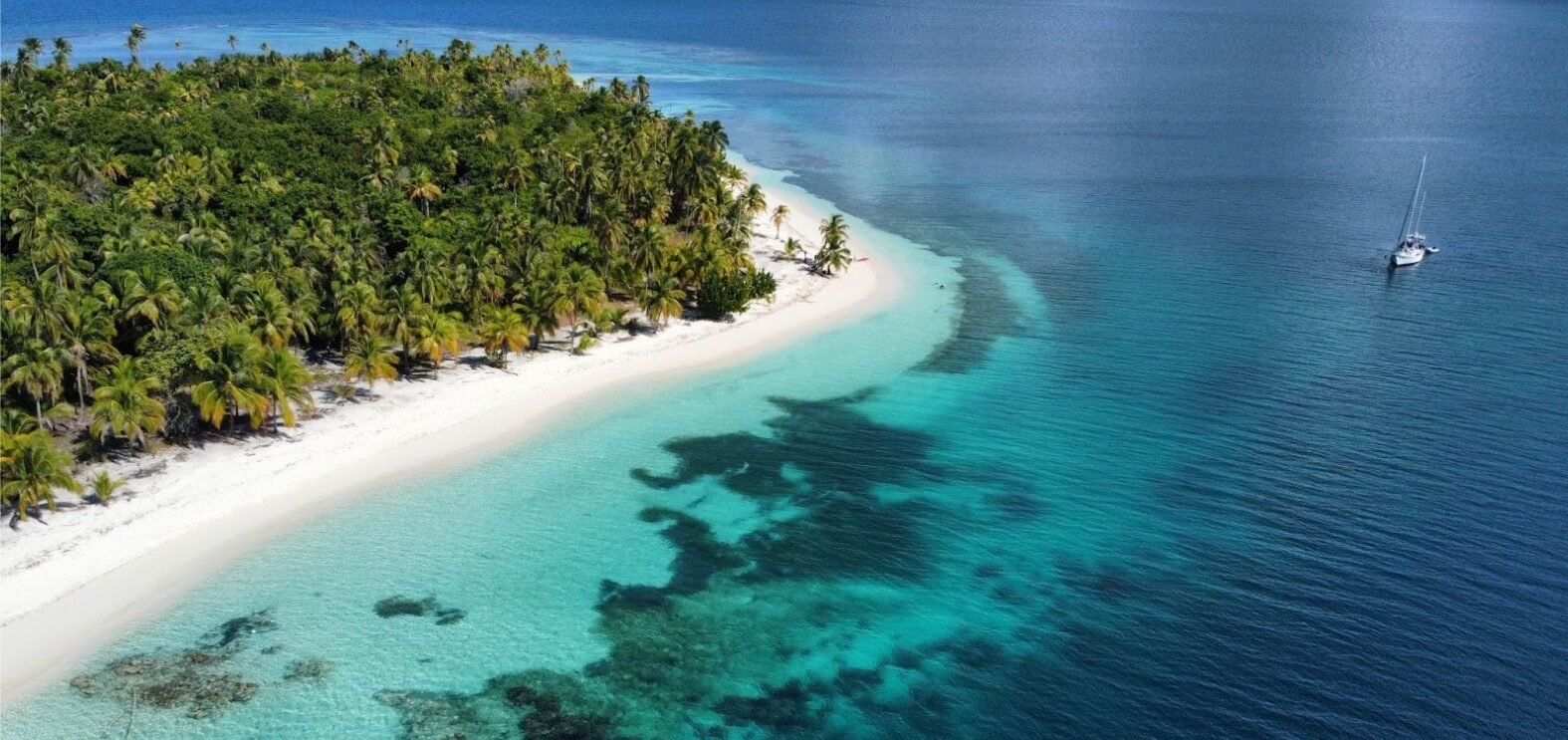 Best Islands to Visit in San Blas, Panama Aerial view of a palm-lined San Blas island with white sand, turquoise water and a sailboat anchored offshore